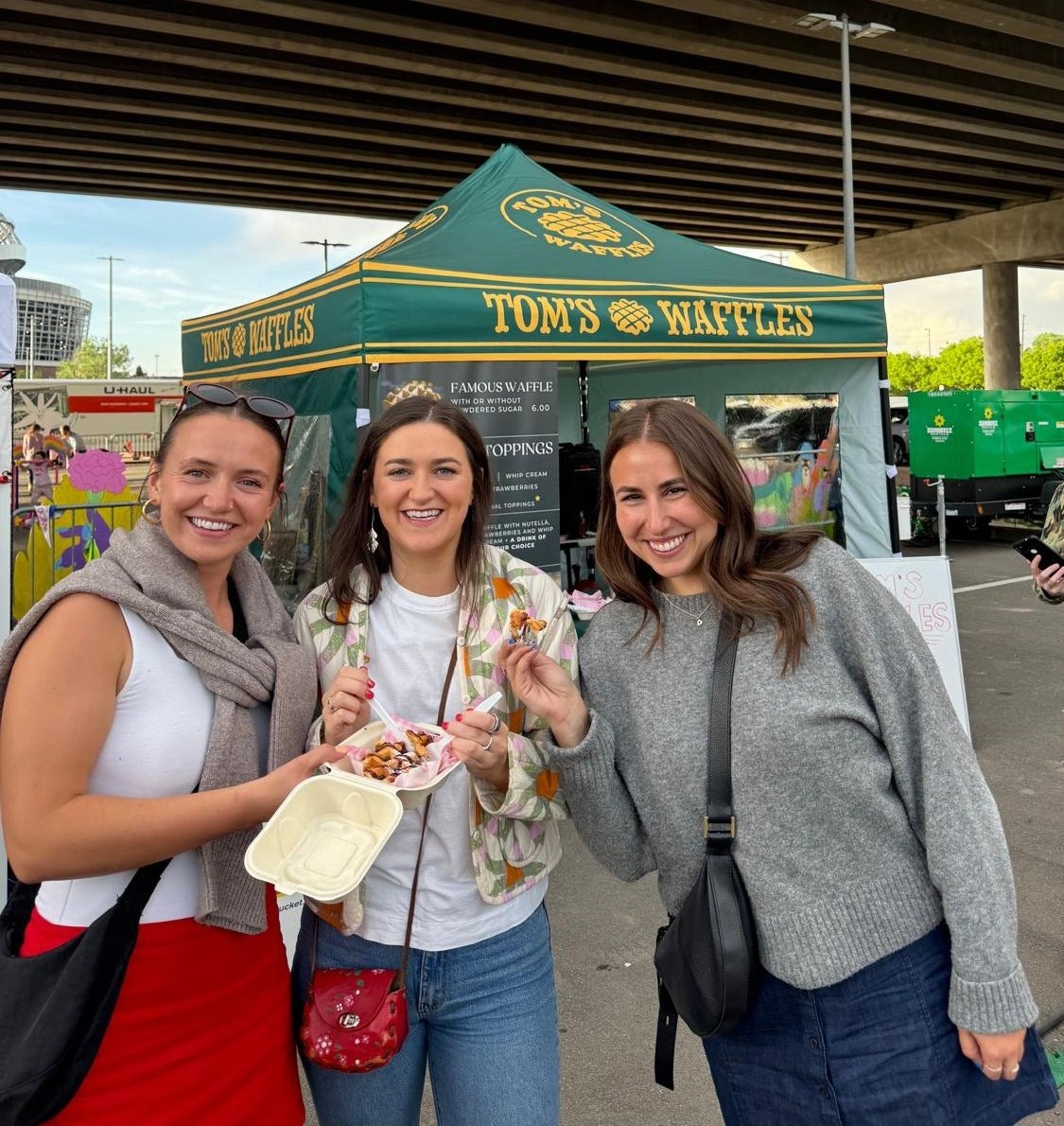 Three guests smiling with a box of waffles in front of the Tom's Waffles tent at a festival