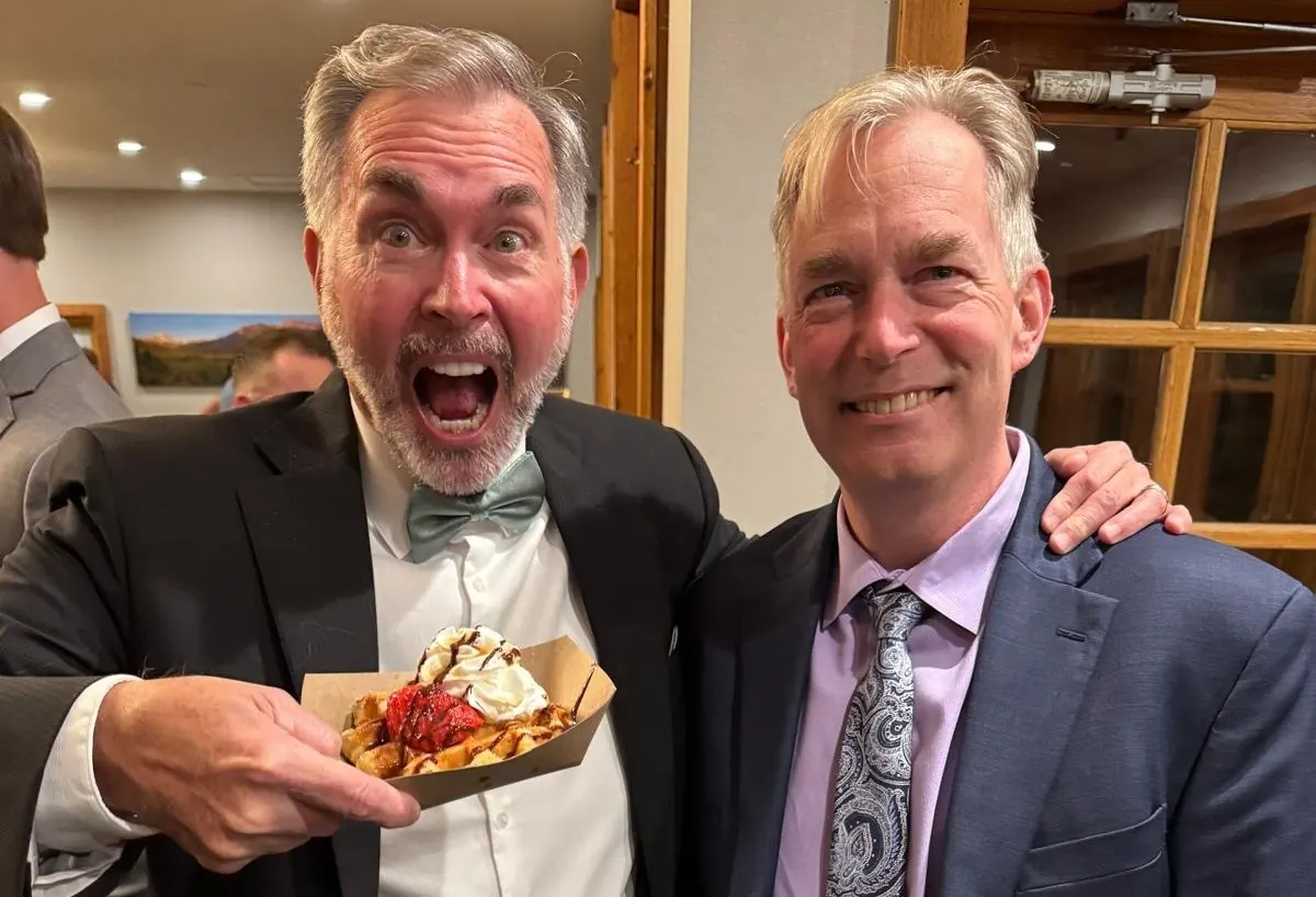 Two men in suits laughing, one holding a waffle with strawberries and cream