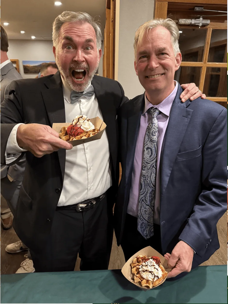 Two men in suits laughing, one holding a waffle with strawberries and cream