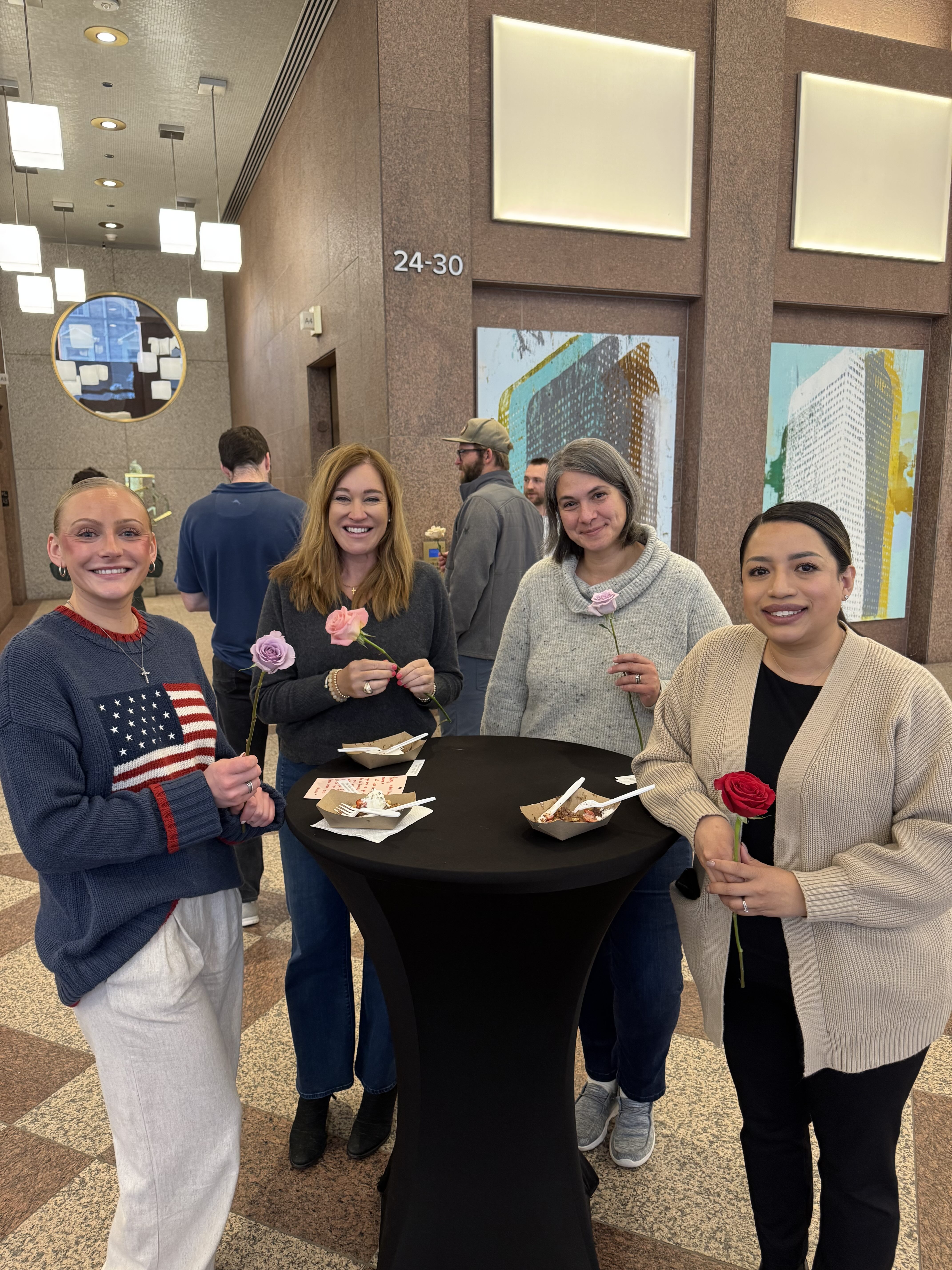 Four coworkers holding roses around a high-top table with their waffles in an office lobby