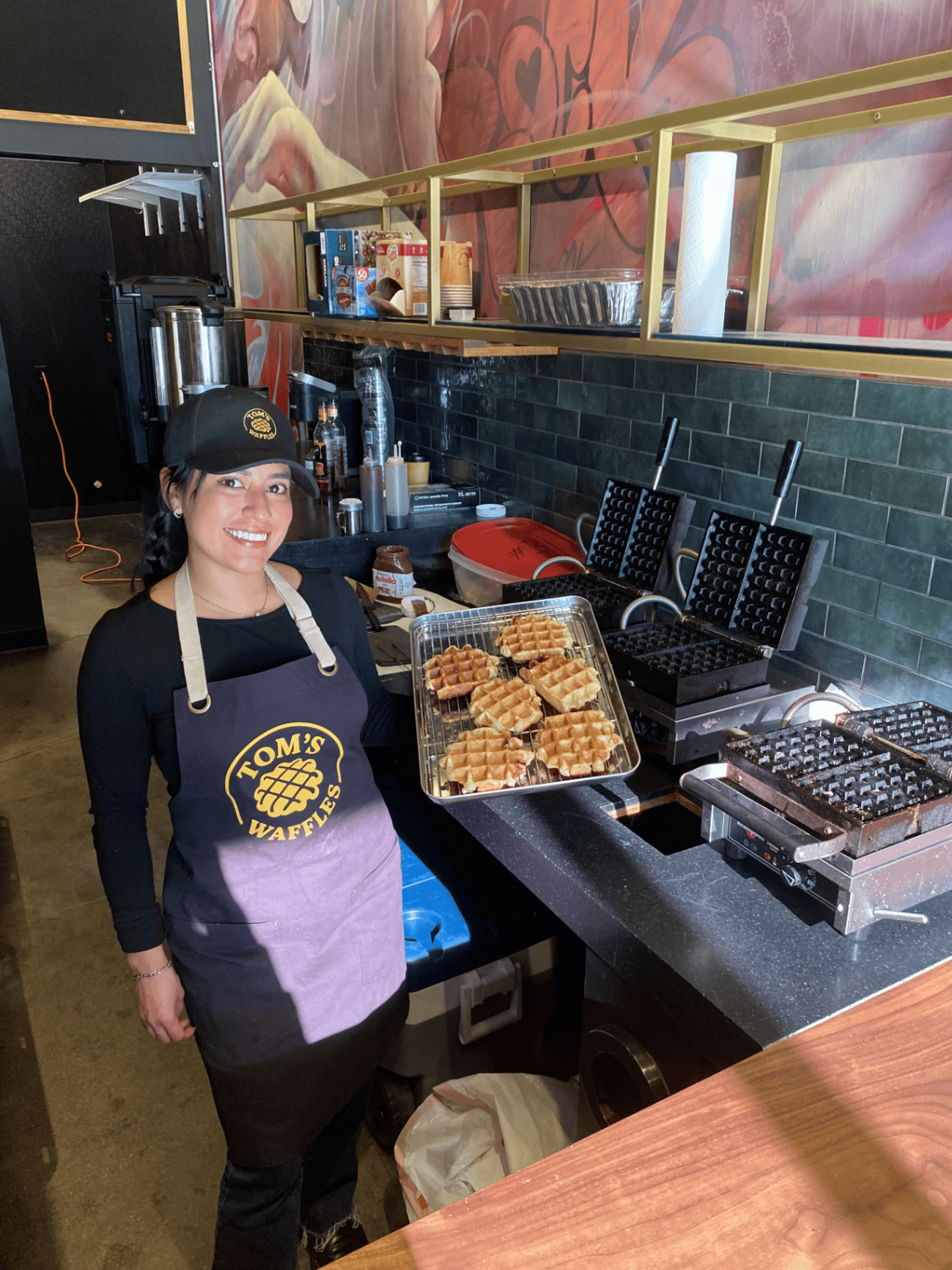 A crew member in a Tom's Waffles apron and cap beside a tray of fresh waffles and the irons
