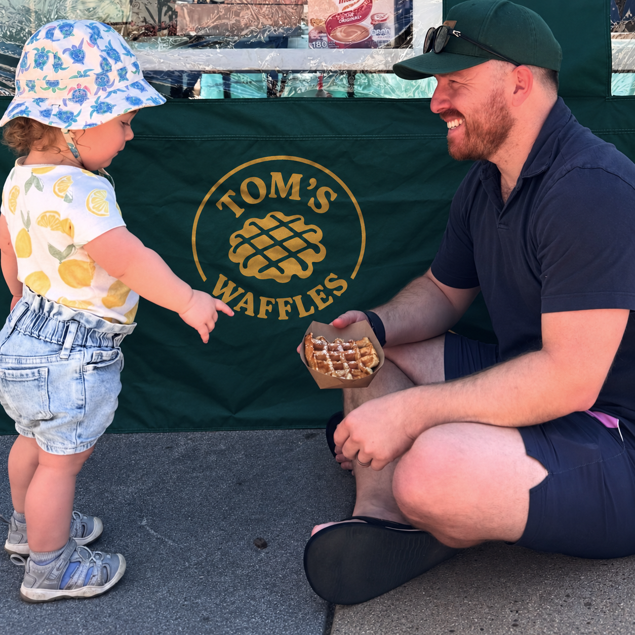 A toddler pointing at a waffle held by a man sitting in front of the Tom's Waffles tent