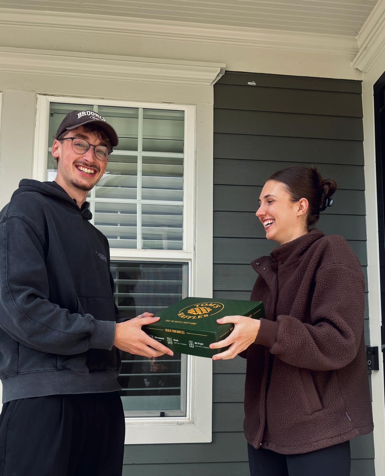 Two people smiling as a Tom's Waffles take-home box changes hands on a front porch